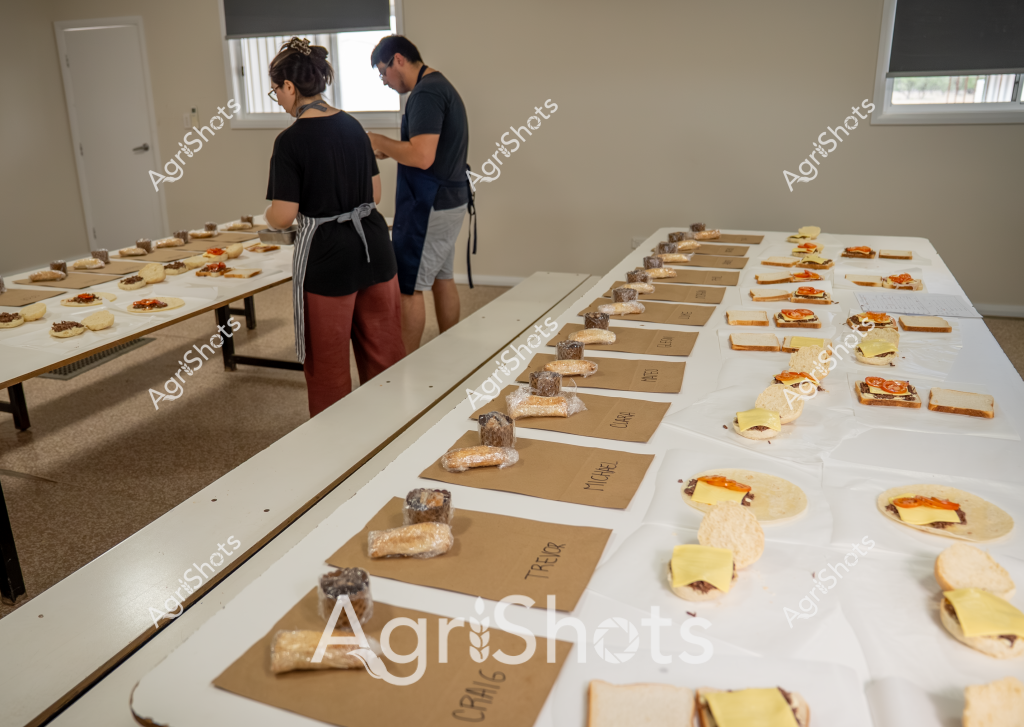 Two people are working on a food preparation and testing setup in an indoor professional kitchen or laboratory environment. A woman and a man, both wearing dark-colored tops and aprons, are arranging multiple food items across several white tables. Kraft paper labels are placed beside various prepared food products - what appear to be sandwiches, pastries, and other prepared dishes.

The scene looks like a systematic food evaluation or sensory testing process, with each item carefully positioned and labeled. Multiple identical food items are arranged in neat rows, suggesting a controlled comparative tasting or recipe development session. The room has neutral beige walls, window blinds, and a professional, organized atmosphere typical of a culinary research or food product development workspace.

The precise arrangement indicates a methodical approach, with each food item positioned with careful consideration. The neutral color palette and clean setup emphasize the professional nature of the food preparation and assessment process taking place.