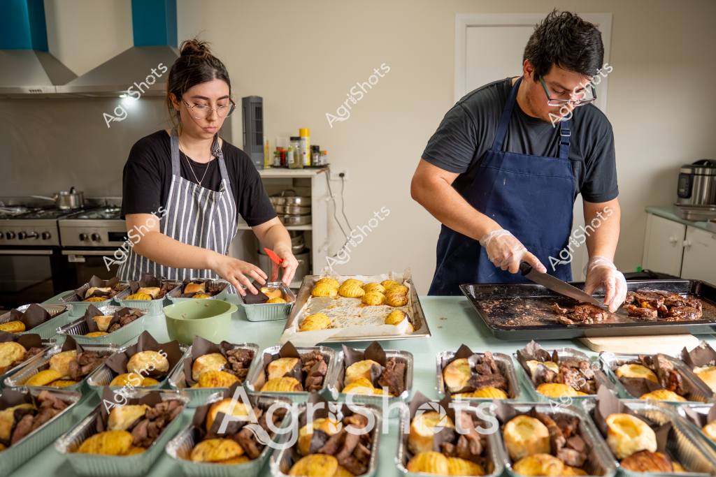 A woman and a man in aprons are meticulously preparing multiple meal portions in a commercial kitchen setting. They are carefully arranging golden-brown baked items and what appears to be beef or meat portions into individual aluminum takeaway containers. The kitchen workspace is filled with numerous prepared meal trays, each consistently featuring a yellow/golden component and a dark meat component. 

The woman, wearing a white and gray striped apron, is using a red tool to adjust items in the containers, while the man in a navy blue apron is cutting or portioning meat on a baking tray. Their focused, precise movements suggest a professional food preparation environment, possibly a catering kitchen or meal prep service. The consistent plating and organized arrangement indicate a systematic approach to batch cooking, with each container featuring a carefully balanced meal composition.

The kitchen environment includes standard stainless steel equipment, cooking utensils, and a neutral color palette, highlighting the methodical and professional nature of their food preparation process. The image captures the behind-the-scenes work of creating multiple identical meal portions with attention to detail and culinary precision.