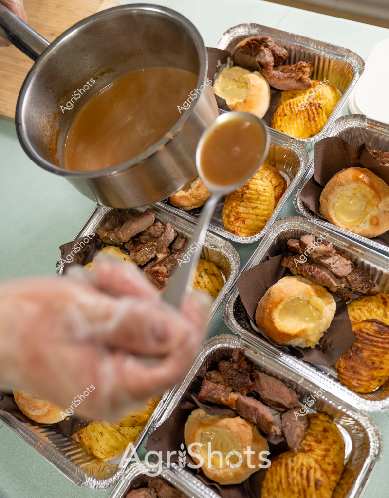 This culinary preparation scene showcases an elegant meat and potato dish being carefully plated in individual aluminum serving containers. A gloved hand is pouring a rich, golden-brown gravy or sauce from a stainless steel saucepan over roasted meat portions and golden, crispy potatoes. The dishes appear to contain tender, browned meat cuts alongside perfectly roasted potatoes with a distinctive ridged or hasselback presentation. Each aluminum container is meticulously arranged, suggesting a professional catering or food preparation environment with careful attention to portion control and presentation. The warm tones of the meat and potatoes contrast beautifully with the metallic surfaces, creating a visually appealing mise en place that hints at a high-quality dining experience.