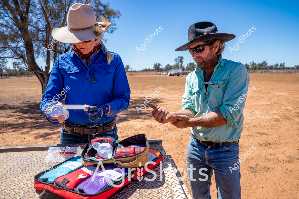 A woman in a wide-brimmed hat and blue long-sleeved shirt and a man in a turquoise shirt and wide-brimmed hat stand beside an open first aid kit on a dusty, sun-baked rural landscape. They appear to be checking medical supplies or preparing emergency equipment, with the arid Australian outback stretching behind them - sparse trees and dry ground indicating a challenging environmental setting. The red first aid kit is open, revealing various medical items and supplies, suggesting they are likely agricultural workers or rural health professionals preparing for potential medical needs in a remote location. The bright blue sky and harsh, open terrain speak to the rugged conditions of Australian rural work environments. Their protective clothing - wide-brimmed hats, long-sleeved shirts, and sturdy work attire - reflects practical preparation for working in intense sunlight and challenging outdoor conditions.
