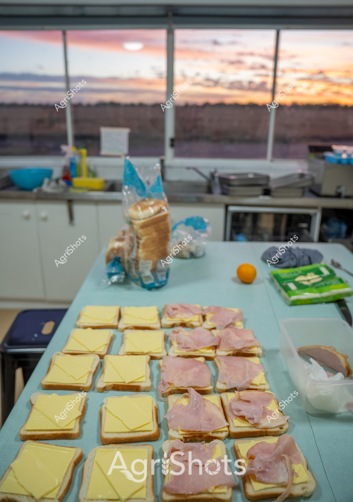 A professional sandwich preparation scene unfolds in a kitchen with a stunning sunset visible through large windows. Multiple slices of bread are meticulously arranged on a blue countertop, with yellow cheese slices and pink ham carefully positioned on alternating pieces, creating a systematic layout for sandwich assembly. A loaf of bread in a partially opened plastic bag sits nearby, along with an orange and other kitchen supplies, suggesting a methodical approach to food preparation. The warm, golden-orange sky outside provides a beautiful backdrop, casting a soft light across the kitchen workspace and creating a serene atmosphere for meal preparation. The precise arrangement of ingredients hints at either commercial food preparation or careful home cooking, with each sandwich component positioned with intentional precision. The scene captures a moment of culinary organization, blending the technical process of sandwich making with the natural beauty of a spectacular sunset.