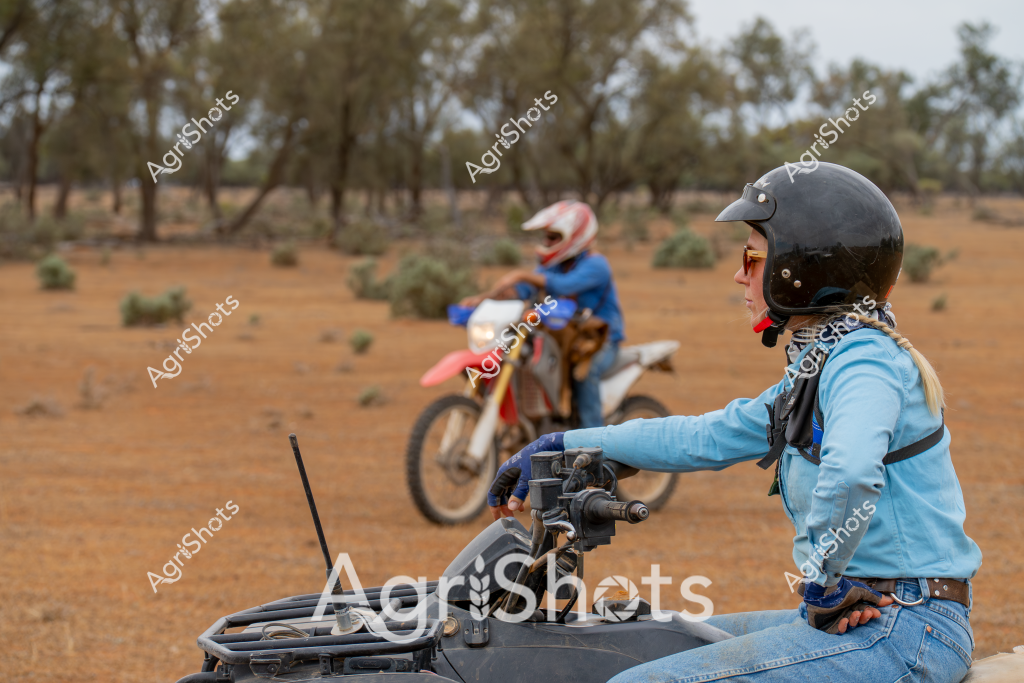 A woman in a blue jacket and black helmet navigates an all-terrain vehicle (ATV) across a dry, open landscape dotted with sparse trees and low shrubs. In the background, another motorcycle rider in a red and blue outfit travels across the dusty, reddish-brown terrain. The scene captures the rugged nature of off-road riding in an arid Australian environment, with the riders demonstrating skill and confidence as they traverse the challenging landscape. The sparse vegetation and open ground suggest this is likely a remote rural or outback setting, where off-road vehicles are essential for traversing extensive properties or exploring challenging terrain. The riders are fully equipped with protective gear, highlighting the importance of safety in these challenging outdoor conditions.