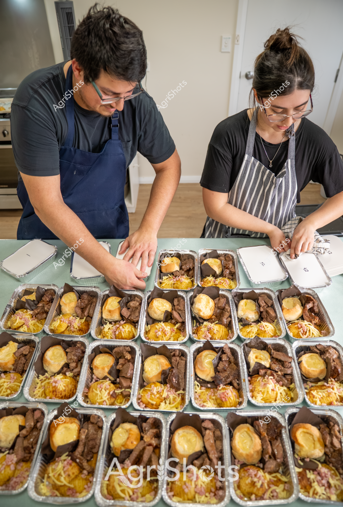 Two individuals in aprons carefully prepare a large batch of meal trays in what appears to be a commercial kitchen or food preparation space. A man wearing a navy blue apron and a woman in a striped gray apron are meticulously arranging multiple aluminum food containers, each filled with a consistent meal composition. The trays contain what looks like beef or meat chunks, golden-topped potatoes or bread rolls, and a bed of yellow-orange shredded ingredients, likely cheese or a similar garnish.

The scene demonstrates precision and coordination, with rows of identically portioned meals systematically laid out across the surface. Their focused demeanor and professional kitchen attire suggest this is likely a commercial food service operation - potentially catering, meal preparation for an institution, or a large-scale food delivery service. The uniform presentation of each tray indicates careful attention to portion control and consistent quality.

The kitchen environment appears clean and organized, with neutral walls and a light-colored work surface. Both individuals are wearing glasses and dark shirts underneath their aprons, and they work side by side with concentrated effort to complete the meal preparation process.
