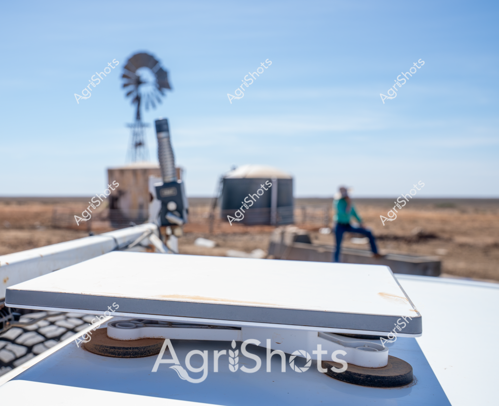 A rural scene captures an agricultural monitoring setup in a dry, expansive landscape, featuring a windmill and technical equipment in the background. In the foreground, a sophisticated white monitoring device with gray trim sits precisely positioned, its sensors and mounting points suggesting advanced data collection capabilities. A person wearing a teal-colored jacket stands near some equipment modules, indicating ongoing field research or environmental monitoring. The stark, arid terrain stretches to the horizon, with a classic Australian windmill standing tall against the clear blue sky, symbolizing traditional water management techniques alongside modern technological approaches. The muted earth tones of the landscape - browns and tans - contrast sharply with the crisp white of the monitoring equipment and the vibrant teal of the worker's clothing, creating a compelling visual narrative of contemporary agricultural science in action.

The scene represents the intersection of traditional farming infrastructure and cutting-edge environmental monitoring technology, typical of agricultural research in Australia's challenging rural environments. The careful placement of equipment suggests precise data collection, potentially related to soil conditions, climate monitoring, or agricultural research.