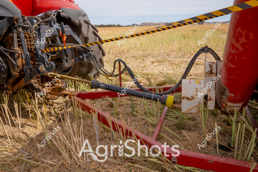 A close-up view of agricultural machinery reveals the intricate connection between a red tractor and its harvesting attachment in a dry stubble field. Hydraulic lines and mechanical linkages connect the farming implement, with yellow and black caution tape stretched across part of the machinery, suggesting recent maintenance or safety protocols. The complex mechanical joints and rugged red paintwork highlight the precision engineering of modern farm equipment. Dried crop stalks are visible beneath the machinery, indicating this is likely during post-harvest season when fields are being prepared or cleaned. The landscape in the background shows a typical Australian rural setting - a flat horizon with distant tree lines and golden-brown grasslands stretching toward the sky, representing the expansive agricultural environments of rural Australia.