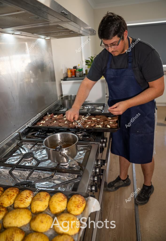 A man in a navy blue apron carefully prepares a tray of roasted meat on a stove, with golden roasted potatoes arranged in the foreground. Standing in a modern kitchen with a stainless steel range hood and gas cooktop, he appears focused on his culinary preparation. The kitchen is clean and well-lit, with cooking ingredients and a small plant visible in the background. His precise movements and professional-looking apron suggest he is carefully tending to the cooking process, arranging what looks like beef or lamb pieces on the baking tray. The golden-brown potatoes in the foreground add a warm, appetizing color to the scene, indicating a carefully prepared meal in progress.