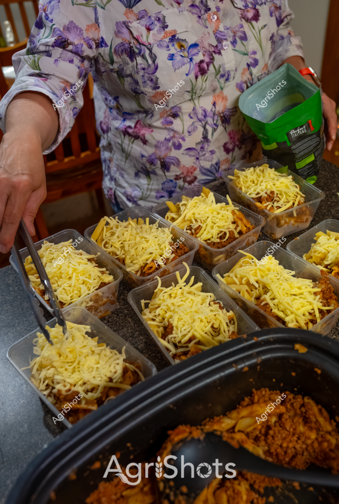A woman in a soft floral blouse is preparing multiple individual meal portions, spreading shredded cheese generously over what appears to be a meat-based dish in small plastic containers. The precise portioning suggests meal preparation or catering, with each container carefully filled with a consistent amount of ground meat and topped with bright yellow grated cheese. A green bottle is visible in her hand, likely a seasoning or sauce to complete the dish. The gray kitchen countertop provides a neutral backdrop to the systematic food preparation, highlighting the methodical approach to creating these individual servings. The cheese appears freshly grated, adding a vibrant yellow color to the meal portions and promising a rich, melted topping when heated.