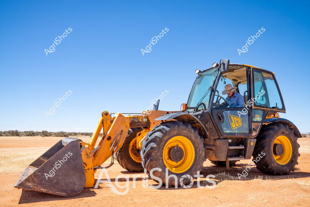 A powerful yellow JCB telehandler sits poised on a dry, dusty agricultural landscape under a brilliant blue sky. The robust machine, with its large distinctive yellow wheels and front-mounted bucket, represents modern agricultural and construction equipment used across Australian rural properties. A man operator sits inside the enclosed cabin, positioned to manage heavy lifting and materials handling tasks typical of farming, construction, and rural infrastructure work. The surrounding environment appears to be an arid or semi-arid region, with sparse vegetation and a wide-open terrain that speaks to the challenging landscapes where such versatile machinery is essential. The telehandler's pristine yellow color contrasts sharply with the terracotta-colored ground, highlighting the machine's industrial strength and purpose in agricultural and rural development settings.
