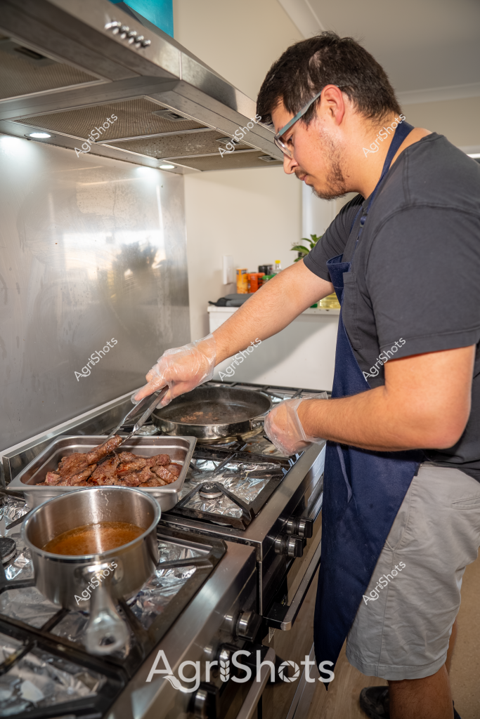 A man in a navy blue apron and dark t-shirt carefully prepares meat in a modern kitchen. He is wearing protective gloves while cooking several pieces of reddish-brown meat in a stainless steel pan on a gas stove. A pot of sauce simmers nearby, and a tray of additional cooked meat sits to the side. The kitchen features a sleek stainless steel rangehood and a reflective backsplash, creating a clean and professional cooking environment. The cook appears focused and precise in his culinary preparation, suggesting a skilled approach to food preparation.