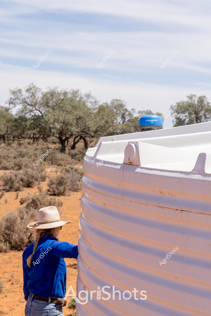 A woman in a blue jacket and wide-brimmed sun hat stands beside a large white water tank in an arid Australian landscape. Sparse eucalyptus trees and low scrubby bushes dot the rusty red earth in the background, typical of a dry inland pastoral setting. The water tank, with a blue top, appears to be a critical water storage resource in this harsh environment. The scene captures the resilience of rural workers managing water infrastructure in a challenging, drought-prone region. The woman's posture suggests careful inspection or maintenance of the water tank, highlighting the importance of water management in Australian agricultural landscapes.