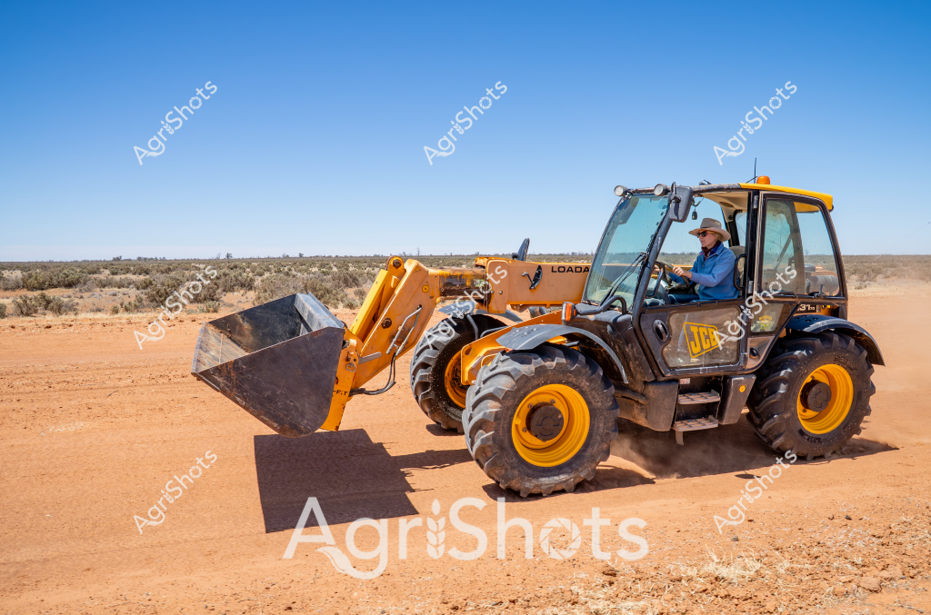 A yellow JCB telehandler operates on a dusty, arid landscape with sparse vegetation in the background. A man wearing a blue shirt and white hard hat carefully maneuvers the heavy machinery across the reddish-brown earth. The telehandler's large front bucket is raised, ready for loading or moving materials, with bright yellow and black paintwork standing out starkly against the clear blue sky. The rugged terrain suggests this is likely a remote agricultural or construction site in the Australian outback, where robust machinery is essential for working in challenging environmental conditions. The JCB's large tires are designed to handle uneven ground, demonstrating the specialized equipment needed to operate in such demanding landscapes. The scene captures the resilience and technological capability required for agricultural and industrial work in Australia's most challenging regions.