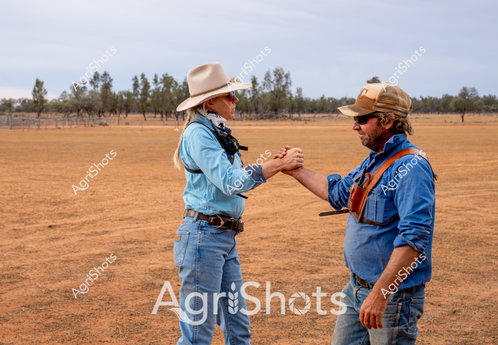 Two people stand in a dry, expansive paddock with scattered trees on the horizon, engaged in a firm handshake. A woman wearing a light blue long-sleeved shirt, light-colored wide-brimmed hat, and blue jeans grips hands with a man in a blue work shirt, brown cap, and denim pants. Their stance suggests a professional agricultural interaction, perhaps concluding a business agreement or land transaction.

The landscape around them is characterized by a parched, ochre-colored ground typical of drought conditions in rural Australia. Sparse eucalyptus trees line the distant background, creating a sense of wide-open pastoral terrain. Both individuals are dressed in practical workwear that reflects the rugged demands of agricultural life, with rolled-up sleeves and sturdy boots suited to the harsh environment.

The handshake symbolizes collaboration and mutual respect in the challenging world of Australian farming, where partnerships and shared understanding are crucial for survival in difficult conditions. Their body language communicates professionalism and a sense of shared purpose against the stark, sun-bleached landscape of what appears to be an inland agricultural property.