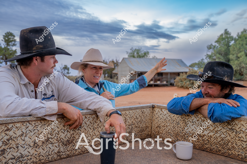 A scene of rural Australian life unfolds at a remote station, with three people in classic stockman attire leaning against a textured metal barrier. Two men and a woman, all wearing wide-brimmed hats and blue work shirts, are engaged in casual conversation with a rustic tin-roofed shed in the background. The landscape suggests an arid or semi-arid region, with reddish earth and sparse trees creating a classic outback setting.

The individuals appear relaxed, with one woman in a light-colored hat gesturing animatedly while speaking to her companions. Their clothing - crisp blue work shirts and sturdy cowboy hats - speaks to the practical nature of station life. A coffee mug sits in the foreground, suggesting a moment of rest during a long working day.

The cloudy sky adds depth to the scene, with soft blue and gray tones creating a moody backdrop to the rural setting. The metal barrier they're leaning on looks weathered, with a diamond-plate texture typical of rural infrastructure. This image captures the essence of Australian rural community - connection, communication, and the shared experience of working the land.

The composition tells a story of camaraderie and the everyday moments that define life on a remote Australian station, where conversation and connection are as important as the work itself.