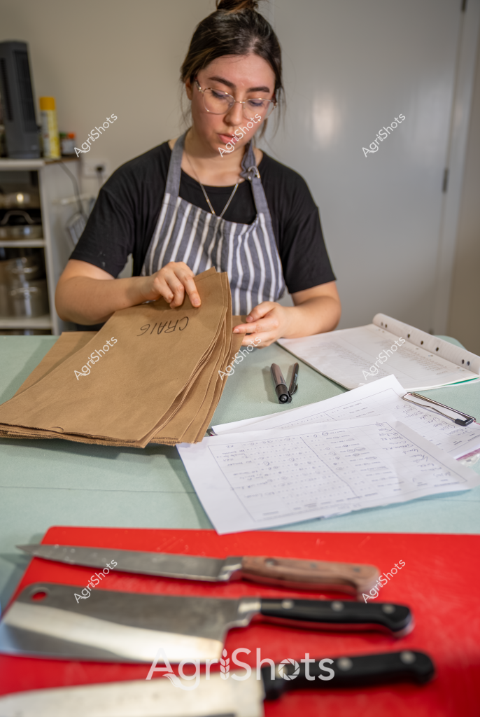 A woman in a striped apron and black shirt carefully handles brown paper bags in what appears to be a professional kitchen or food preparation environment. She is wearing glasses and has her hair pulled back, focusing intently on her task at a workstation. Several professional kitchen knives are arranged on a red cutting board in the foreground, suggesting a culinary workplace. White papers and documents are spread across the work surface, indicating an organized and methodical approach to food preparation or packaging. The setting looks clean and professional, with neutral-colored walls and kitchen equipment visible in the background, reflecting a structured and hygiene-conscious workspace.
