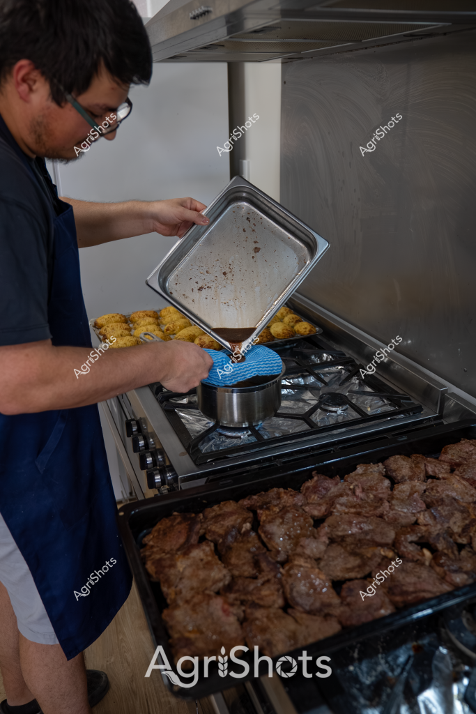 A man in a navy blue shirt carefully tilts a metal tray over a gas stovetop, pouring what appears to be cooking drippings or sauce into a small pot. The kitchen scene reveals multiple cooking stages, with golden-brown breaded items on one tray and browned meat pieces on another baking tray. He is wearing glasses and using a blue silicone mitt to handle the hot metal tray, suggesting a careful and methodical approach to food preparation. The stainless steel stove and overhead exhaust fan indicate a professional or well-equipped home kitchen setting, with multiple dishes in various stages of cooking or resting. The image captures a moment of culinary precision, with the man focused on transferring liquid from one cooking vessel to another.