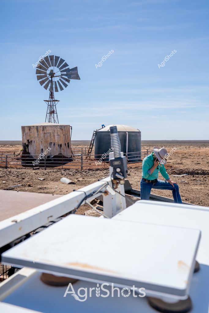 A lone figure in a turquoise shirt and wide-brimmed hat sits contemplatively beside a classic Australian windmill in an expansive, arid landscape. The rusted metal windmill stands tall next to weathered water tanks, surrounded by a barren, rocky terrain that epitomizes the challenging conditions of outback farming. A white equipment surface dominates the foreground, suggesting this is a working agricultural site where water infrastructure is critical for survival in a dry environment. The clear blue sky stretches endlessly, emphasizing the isolation and resilience required in remote agricultural settings. Water storage tanks and the iconic windmill represent the essential infrastructure that enables farming and livestock management in Australia's harsh, drought-prone regions. The scene captures the solitude and quiet determination of rural workers who maintain agricultural operations in extremely challenging environments.