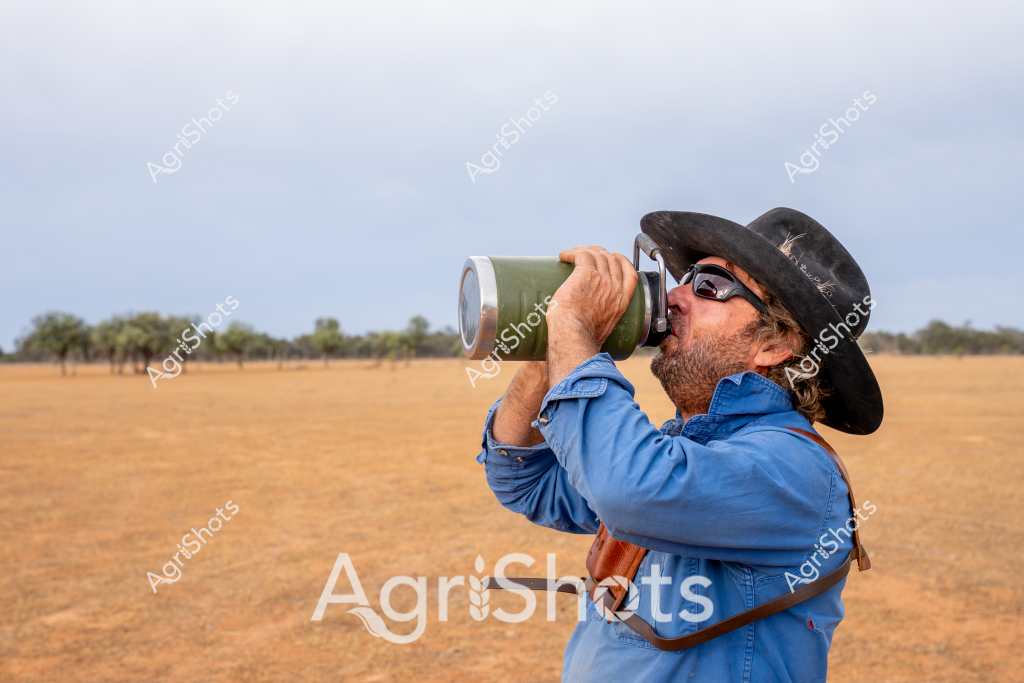 A man in a blue denim shirt and black wide-brimmed hat drinks intently from a large green water bottle while standing in a dry, golden grassland. The rugged landscape stretches out behind him, with scattered trees on the distant horizon and a pale, overcast sky creating a sense of vast, arid Australian countryside. Wearing sunglasses and displaying a weathered, unshaven appearance, the individual seems prepared for the harsh rural environment, embodying the resilient spirit of outback workers. His posture suggests he is taking a crucial water break during what appears to be a long, hot day of agricultural work. The parched field and his intense drinking pose capture the challenging conditions faced by rural workers in drought-affected regions.