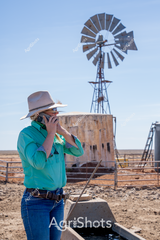 A woman in a teal work shirt and wide-brimmed hat stands amid a dry, arid rural landscape, talking on a mobile phone near a classic Australian windmill and water tank. The rural setting captures the essence of remote station life, with a metal windmill towering against a clear blue sky and a concrete water trough in the foreground. Her outfit - a bright turquoise long-sleeved shirt, blue jeans, and a neutral-toned rural work hat - speaks to practical station wear for harsh outback conditions. The windmill, a quintessential piece of Australian agricultural infrastructure, stands tall on a metal tower, its multiple metal blades designed to pump groundwater in this seemingly dry, rocky terrain. The landscape suggests a remote farming or cattle station location, likely in Western Australia, Northern Territory, or Queensland, where water infrastructure is critical for livestock and agricultural survival.