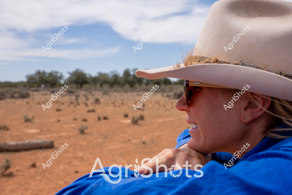 A woman wearing a light-colored wide-brimmed cowboy hat and sunglasses looks out across a sun-baked, rust-colored Australian outback landscape. She is dressed in a bright blue top and appears contemplative, surveying the arid terrain with its scattered sparse vegetation and distant trees. The sky is a soft blue with wispy clouds, creating a sense of expansive openness typical of remote rural Australian settings. Her profile suggests a calm, experienced demeanor of someone accustomed to working in challenging agricultural environments. The image captures a moment of quiet observation, reflecting the resilience and connection to land that characterizes rural Australian life.