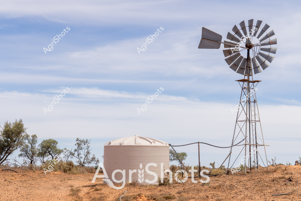 A classic Australian rural landscape captures a weathered windmill and water tank in an arid, drought-affected setting. The tall metal windmill stands prominently against a soft blue sky, its metal blades positioned to catch any available breeze. A corrugated steel water tank sits nearby, providing crucial water storage for the surrounding sparse landscape. Scattered native scrub and sparse trees dot the reddish-brown earth, highlighting the challenging environmental conditions typical of inland Australia. The scene embodies the resilience of rural infrastructure, with the windmill representing generations of agricultural adaptation to harsh, dry environments. The muted color palette of dusty browns, soft blues, and silvery metal tones speaks to the austere beauty of Australia's agricultural heartland.