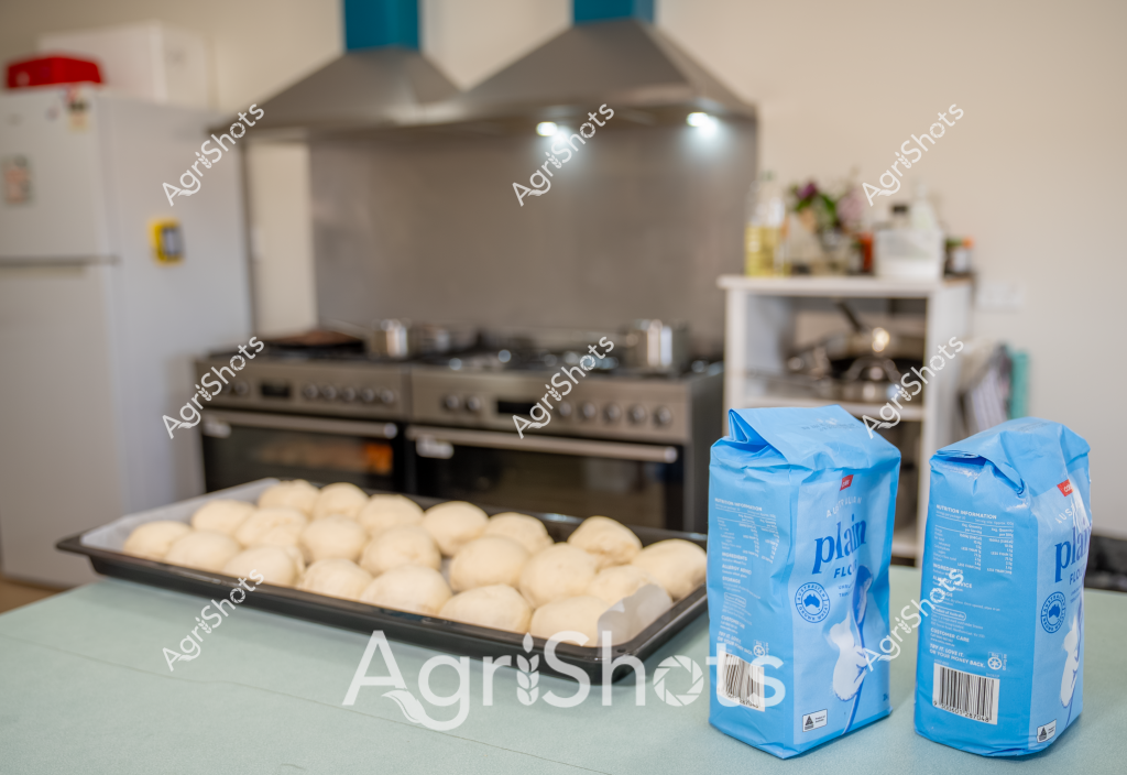 Based on the image, here's a detailed description:

A kitchen scene captures the early stages of bread making, with rows of perfectly shaped dough balls resting on a baking tray. Two blue bags of plain flour sit in the foreground, suggesting home baking or small-scale bakery preparation. In the background, a professional stainless steel stove and range hood create a clean, professional cooking environment. The raw dough appears uniform and carefully shaped, indicating skilled preparation - each round ball is smooth and nearly identical, waiting to be transformed by the oven's heat. The soft cream-colored dough contrasts beautifully with the cool blue flour packaging and the sleek metallic kitchen equipment, creating a sense of culinary anticipation and precision. The image captures a moment of potential, where simple ingredients are about to become something delicious, showcasing the careful craft of bread making.