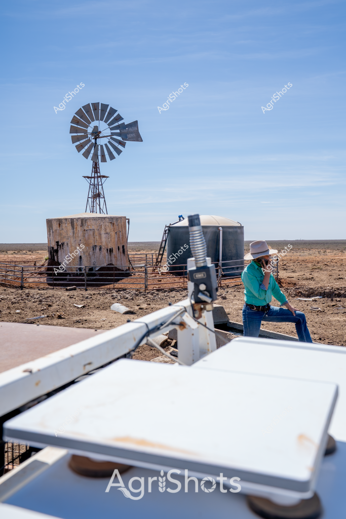 A windmill and water storage setup dominates this arid rural landscape, capturing the essence of remote Australian farming infrastructure. A person wearing a turquoise shirt and a wide-brimmed hat sits contemplatively near water tanks, surrounded by a stark, dusty terrain that stretches to a flat horizon under a clear blue sky. An old metal windmill stands prominently, its multiple blades positioned to catch even the slightest breeze, alongside a weathered concrete water storage tank enclosed by metal fencing. The scene epitomizes the resilience of agricultural operations in harsh, dry environments, where water management is critical for survival. Rusted equipment and minimal vegetation underscore the challenging conditions of this remote agricultural setting, highlighting the technological adaptations farmers use to sustain operations in Australia's unforgiving outback landscape.