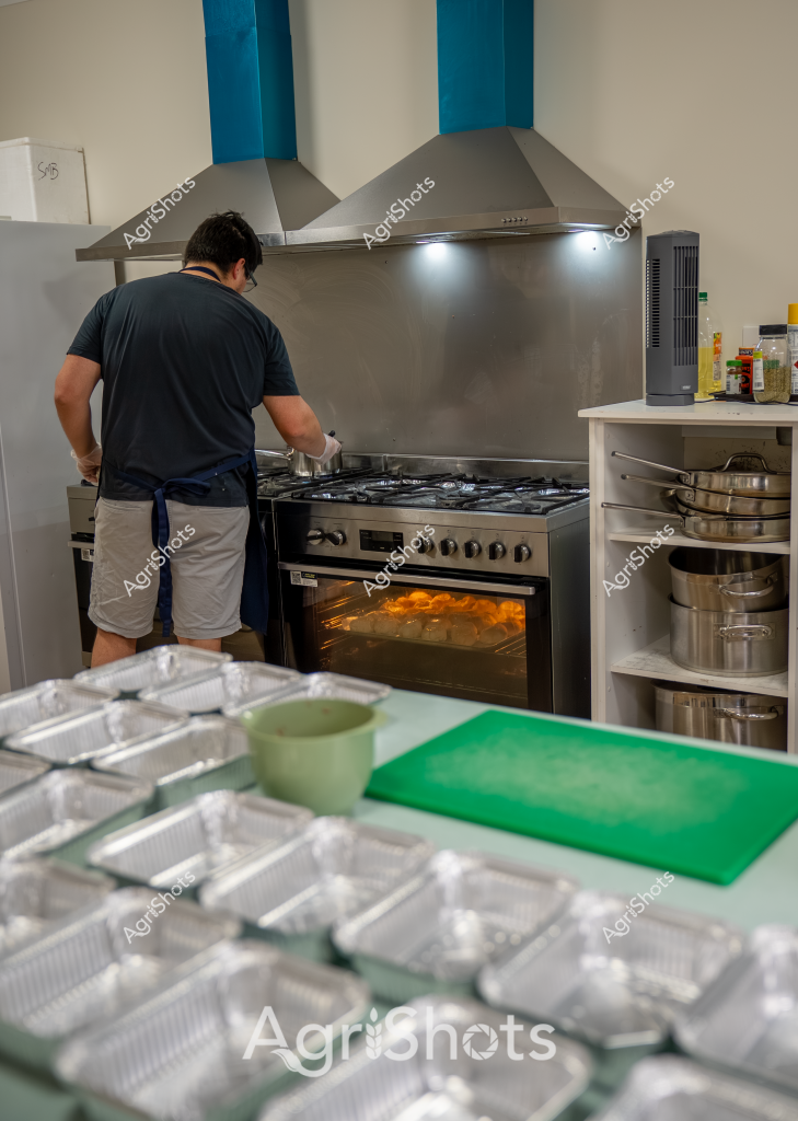 A man in a dark shirt and apron works diligently in a commercial kitchen, preparing multiple meal portions in disposable aluminum containers. The stainless steel kitchen features a professional range with an oven emitting a warm, golden glow, suggesting food is currently being cooked. Numerous identical aluminum food trays are arranged on a green and white preparation surface, indicating a systematic approach to food production, likely for catering, meal delivery, or institutional food service. Stainless steel pots are neatly stacked on shelves to the right, and industrial-grade range hoods provide ventilation above the cooking area. The scene captures a moment of focused culinary preparation, with precision and efficiency evident in the organized workspace.