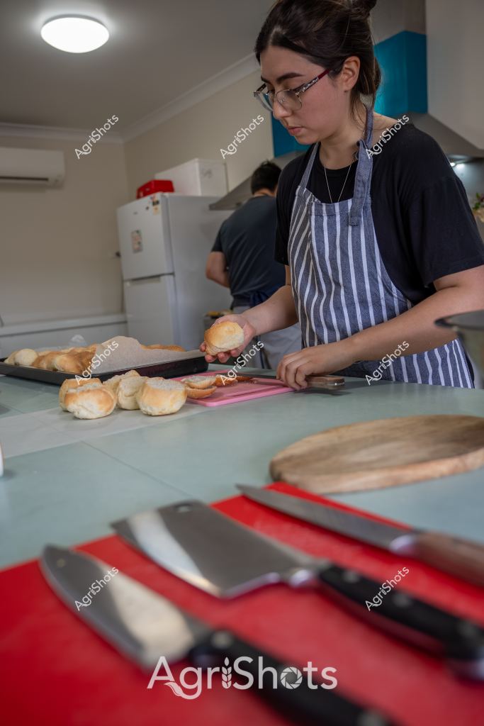 A young woman wearing glasses and a blue-and-white striped kitchen apron prepares bread rolls in a commercial kitchen setting. She stands at a work surface with several golden-brown bread rolls, carefully handling one while other rolls are arranged on a baking tray. In the foreground, professional kitchen knives are laid out on a red cutting board, suggesting a professional food preparation environment. The kitchen appears clean and well-lit, with a white refrigerator visible in the background and another person partially seen working nearby. The woman demonstrates focused concentration as she works, her precise movements indicating skill and attention to detail in food preparation.