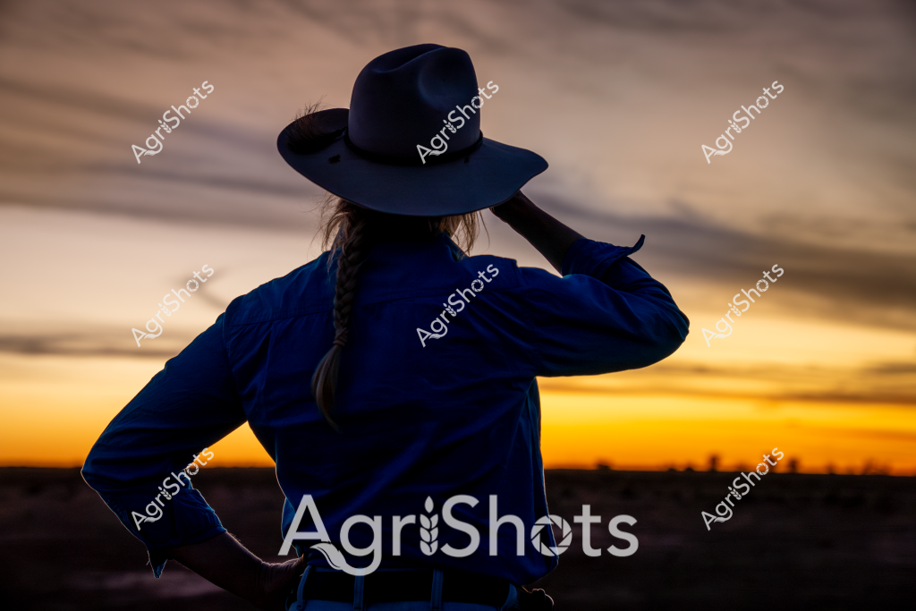 A woman wearing a wide-brimmed dark blue hat stands with her back to the camera, silhouetted against a stunning sunset landscape. Her blue jacket and braided hair are clearly visible as she looks out across the golden horizon, her hand adjusting her hat. The sky blazes with warm orange and yellow tones, creating a dramatic backdrop that stretches across a rural landscape. The scene captures a moment of quiet contemplation, with the figure seeming to pause and take in the breathtaking evening light of what appears to be an Australian rural setting. The interplay of deep blue clothing against the vibrant sunset creates a powerful visual contrast, evoking the sense of connection between a person and the expansive agricultural landscape.
