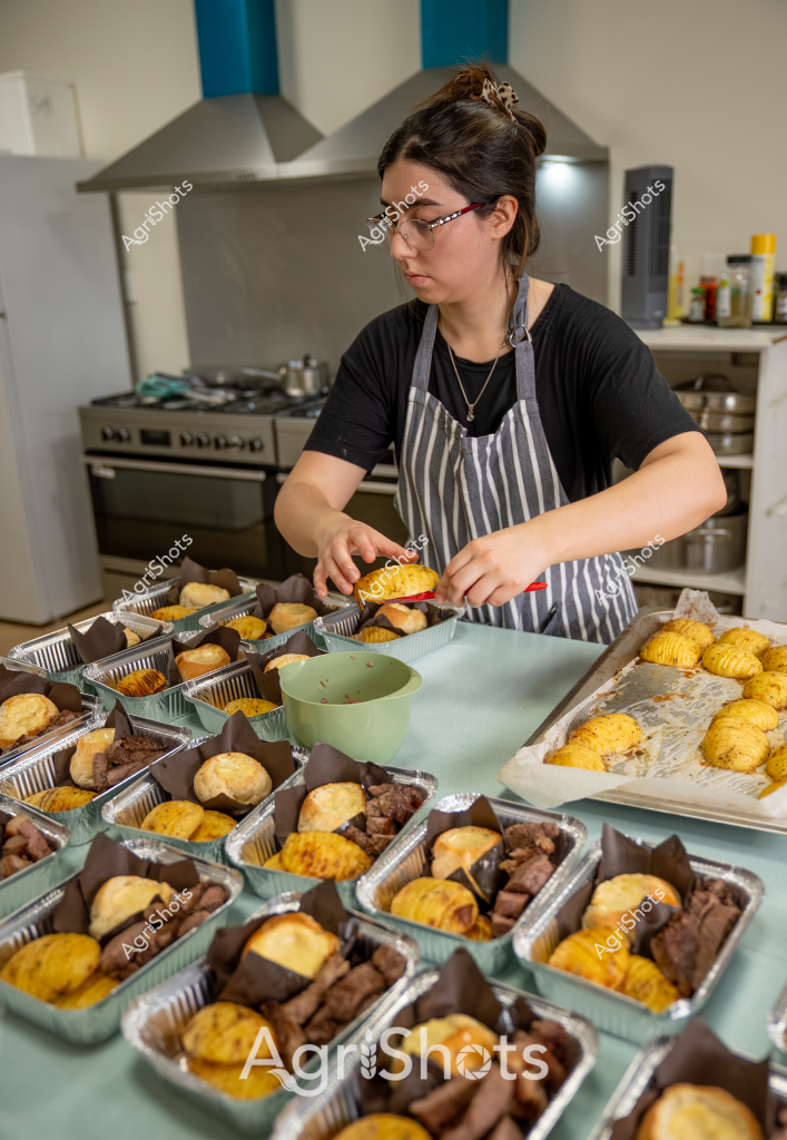 A woman wearing a striped apron and black shirt is meticulously preparing food portions in an industrial kitchen. Multiple aluminum trays are arranged across the workspace, each containing yellow baked items and what appear to be dark brown protein pieces. She is carefully positioning and organizing these pre-portioned meal components, demonstrating precision and attention to detail in her food preparation process. The kitchen environment features a stainless steel stove and range hood in the background, with cooking ingredients and equipment visible on surrounding surfaces. The uniform arrangement of the meal trays suggests this is likely a catering or commercial food service preparation setting, with each tray containing a consistent combination of golden and brown food items.
