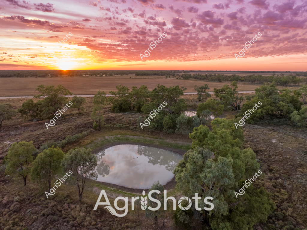 A stunning aerial landscape capturing a serene rural waterhole surrounded by Australian bushland during a spectacular sunset. The small, circular water dam is nestled among scattered eucalyptus trees, reflecting the dramatic pink and orange sky with soft cloud formations. Surrounding the waterhole are dry, rugged paddocks and sparse vegetation typical of an Australian agricultural landscape, with cultivated fields visible in the distant background. The golden sunlight breaks across the horizon, casting a warm glow over the terrain and highlighting the intricate textures of the landscape. This image beautifully illustrates the critical importance of water infrastructure in rural and agricultural settings, showcasing how strategic water management supports ecosystem survival in arid and semi-arid regions. The natural symmetry of the waterhole, encircled by green trees against the earthy brown landscape, creates a compelling visual representation of Australia's resilient agricultural environments.
