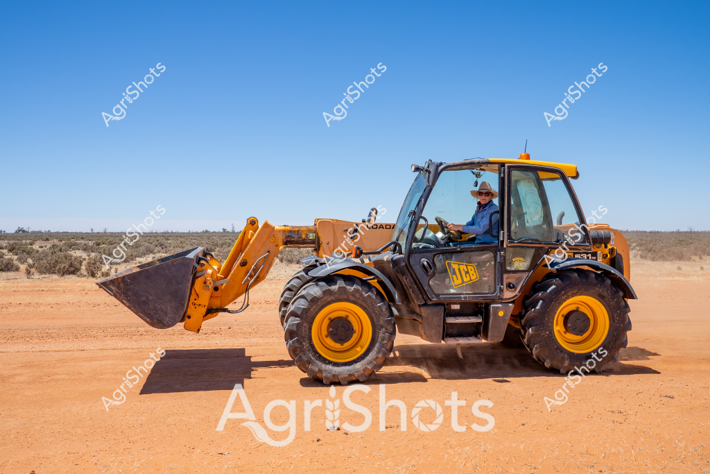 A JCB telehandler stands ready in a sun-drenched, arid landscape, its vibrant yellow and black color scheme contrasting sharply against the rich terracotta-colored dirt and sparse scrubland. A man wearing work attire and a broad-brimmed hat operates the heavy machinery, positioning the large front bucket against a clear blue sky. The robust agricultural vehicle features substantial chunky tyres designed for navigating challenging rural terrain, with the distinctive JCB branding prominently displayed on its side panel.

The telehandler appears well-maintained and prepared for agricultural or construction work, typical of machinery used in remote Australian farming and resource extraction regions. Its muscular profile suggests powerful lifting capabilities, essential for tasks like moving materials, loading supplies, or supporting infrastructure development in challenging outback environments.

The surrounding landscape hints at a remote location - possibly in Western Australia, South Australia, or the Northern Territory - with low scrub vegetation and a vast, open horizon stretching into the distance. The harsh, bright lighting and cloudless sky indicate midday conditions in a hot, dry agricultural or mining region.