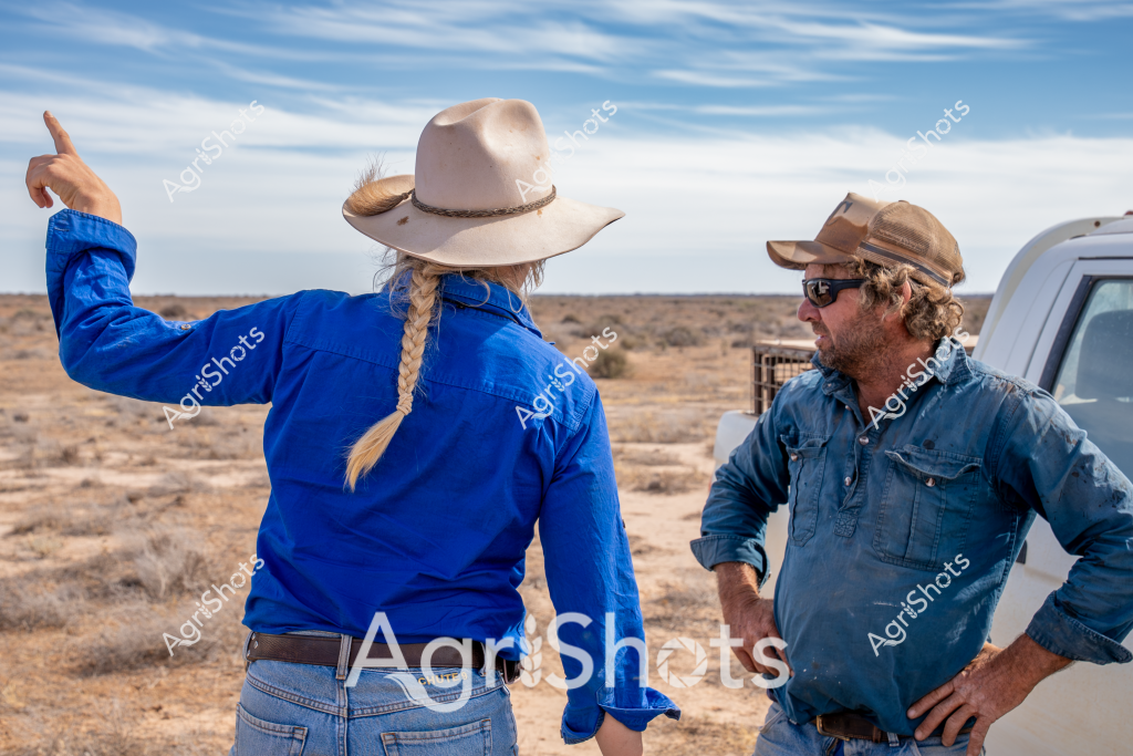 Two people stand in a dry, sparse desert landscape, discussing their work. A woman wearing a blue long-sleeved shirt and a cream-colored wide-brimmed hat faces away from the camera, her blonde braid hanging down her back. She points with one arm while gesturing to something in the distance. Beside her, a man in a denim shirt and a brown cap with sunglasses stands with his hand on his hip, listening intently. A white utility vehicle is parked nearby, suggesting they are likely agricultural workers or station managers surveying a remote property. The landscape is characterized by low scrubby vegetation, sandy ground, and a vast open sky with wispy clouds, typical of Australia's arid interior. The scene captures the collaborative nature of rural work, with both individuals engaged in what appears to be a serious discussion about the surrounding terrain or a specific task at hand.