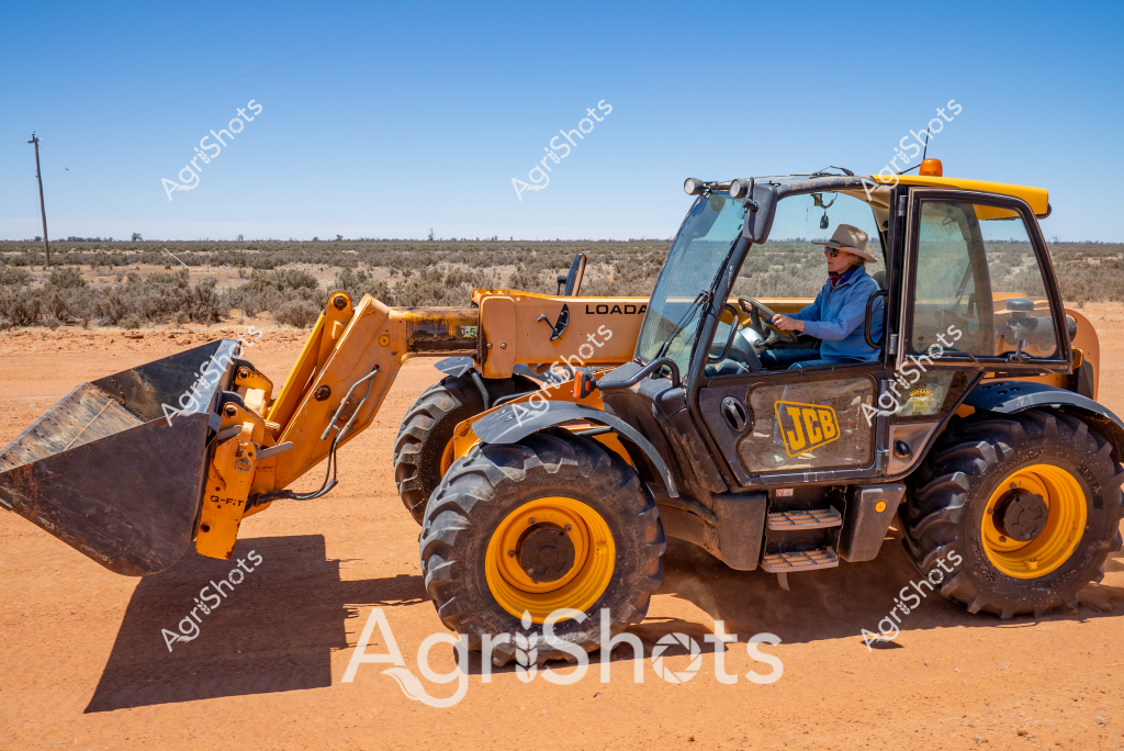 A man operator sits in a bright yellow JCB telehandler (telescopic loader) against a stark, arid Australian landscape. The heavy-duty agricultural machine stands on deep red dirt with sparse scrubland stretching to the distant horizon, typical of Australia's interior regions. The telehandler features robust yellow and black color styling, with large chunky wheels designed for rough terrain and a substantial front-mounted loading bucket. The operator, wearing a light blue shirt and a wide-brimmed hat for sun protection, is positioned inside the air-conditioned cabin, suggesting this is likely a hot, dry day in an outback or remote agricultural setting. The machine appears ready for earthmoving, construction, or farm infrastructure maintenance tasks, highlighting the critical role of specialized equipment in challenging Australian rural environments.