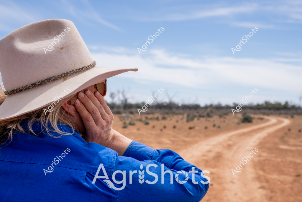 A woman wearing a wide-brimmed cream-colored hat and a bright blue shirt gazes across a dusty, arid landscape. Her hand rests against her face as she looks out over a dry, reddish-brown dirt track winding through sparse scrubland. The sky is a soft blue with wispy clouds, creating a sense of quiet contemplation and the vastness of the Australian outback. Her pose suggests a moment of reflection, perhaps considering the challenging rural environment or the day's agricultural work ahead. The landscape speaks to the resilience of rural workers and the harsh beauty of Australia's interior regions, where every vista tells a story of survival and connection to the land.