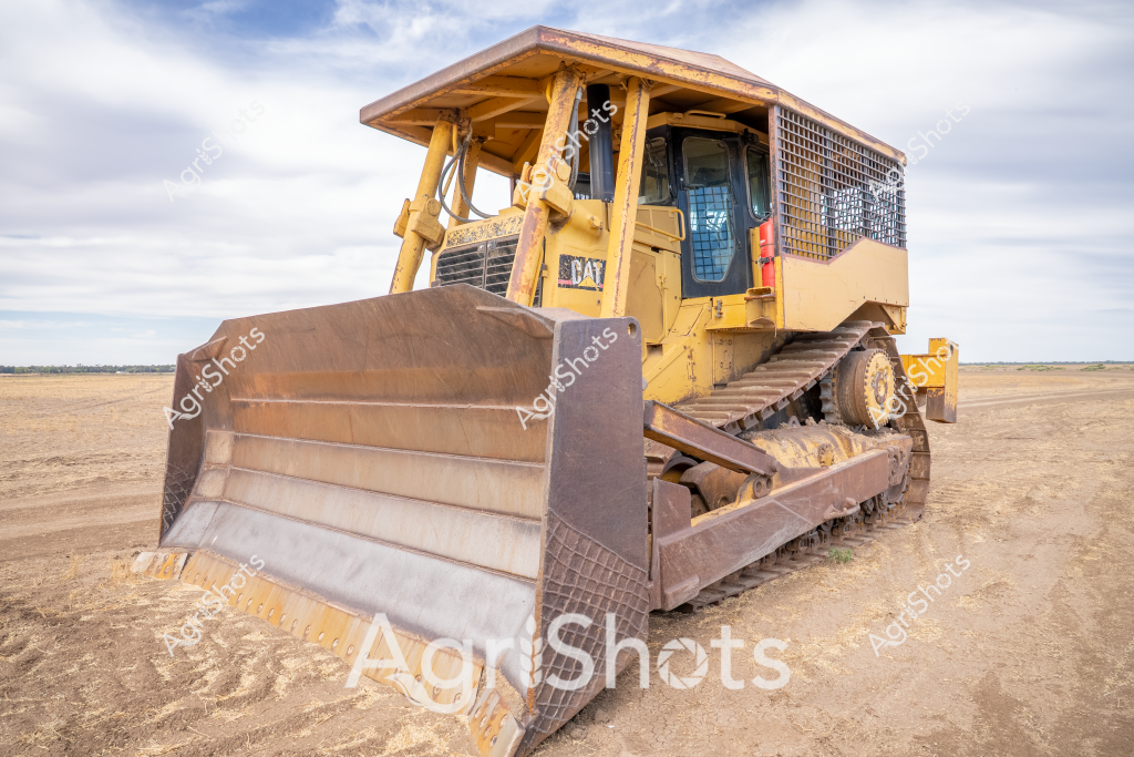 A yellow Caterpillar (CAT) bulldozer sits on a dry, dusty landscape, showcasing the robust machinery used in Australian agricultural and construction operations. The heavy-duty tracked vehicle features a large metal blade at the front, designed for earth-moving, land clearing, and site preparation tasks. Its weathered yellow paint and sturdy construction reflect the demanding conditions of rural and industrial environments. The expansive flat terrain stretches to the horizon, with a cloudy sky providing a muted backdrop that emphasizes the machine's significant presence. The bulldozer's protective cabin and mesh safety guards highlight the engineering considerations for operator protection in challenging work settings. This powerful piece of equipment represents the critical role of heavy machinery in developing and maintaining agricultural and construction landscapes across Australia.