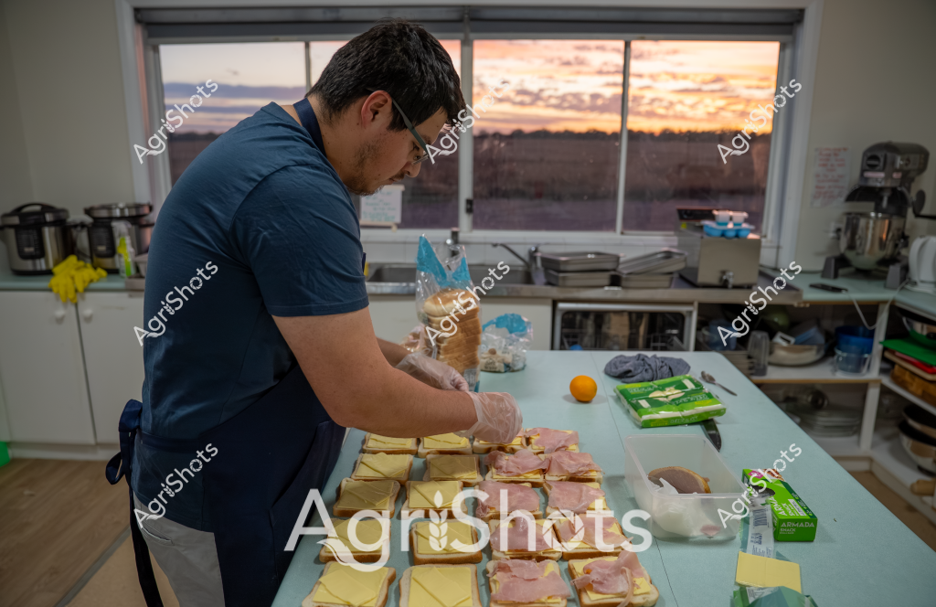 A professional preparing sandwiches in a commercial kitchen, with a stunning sunset visible through large windows behind him. The man, wearing a navy blue shirt and food preparation gloves, is carefully assembling multiple sandwich portions on a clean work surface. Slices of yellow cheese and pink ham are meticulously laid out across several bread bases, suggesting a systematic food preparation process. Kitchen equipment like industrial mixers and appliances are visible in the background, while a warm, golden sunset casts a beautiful glow through the windows, creating a serene atmosphere in the workspace. An orange sits nearby, and green packaging suggests a professional catering or food service environment.