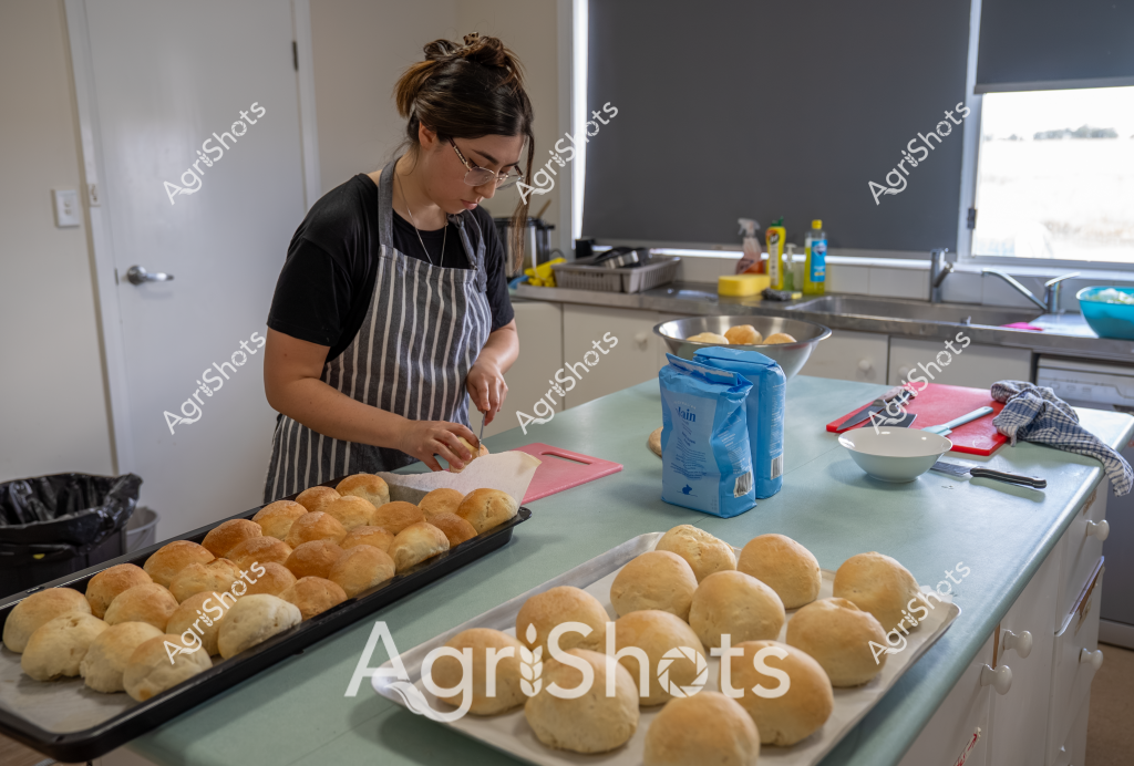 A woman baker in a striped apron and black t-shirt meticulously prepares a batch of golden bread rolls in a modern kitchen. Multiple trays are filled with beautifully risen, uniformly shaped bread rolls, displaying a consistent golden-brown color and smooth surface. A bag of flour sits nearby on the kitchen counter, along with baking tools and a pink cutting board, suggesting she is in the midst of a careful bread-making process. Soft natural light filters through a window blind, creating a calm and focused atmosphere in the clean, contemporary kitchen space. The precise arrangement of the bread rolls and the baker's concentrated posture indicate professional-level baking skills and attention to detail in her craft.