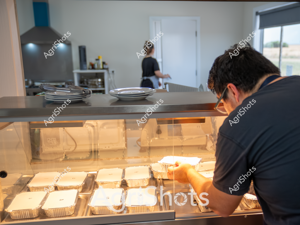 A professional kitchen scene captures a worker preparing pre-packaged meals in aluminium trays. A person wearing a dark shirt is carefully arranging multiple uniform food containers in a heated display cabinet, with stainless steel surfaces and kitchen equipment visible in the background. The precise arrangement of identical aluminium food trays suggests a commercial food preparation environment, likely for catering, meal delivery, or institutional food service. Warm lighting illuminates the gleaming metal surfaces and packaged meals, highlighting the systematic and hygienic approach to food preparation. Another person can be seen in the background working in the kitchen, indicating a busy, collaborative workspace dedicated to efficient meal production.