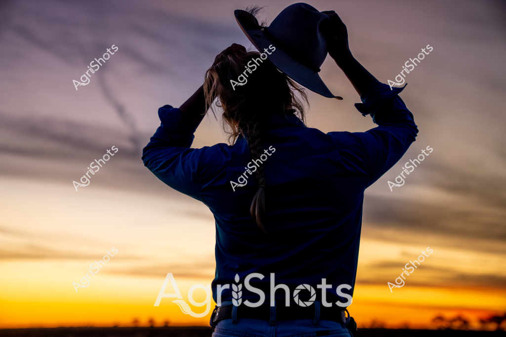 A woman in a blue jacket stands with her back to the camera, silhouetted against a stunning sunset sky with vibrant yellow and orange hues blending into deep purple clouds. She is holding a wide-brimmed hat, with her hair visible and slightly windblown, suggesting an outdoor rural setting. The dramatic twilight landscape stretches out behind her, with shadowy tree lines marking the distant horizon. The scene captures a contemplative moment, representing the quiet transition between day and evening in an expansive agricultural landscape. The rich, warm colors and the solitary figure evoke a sense of peaceful reflection and connection to the natural environment.