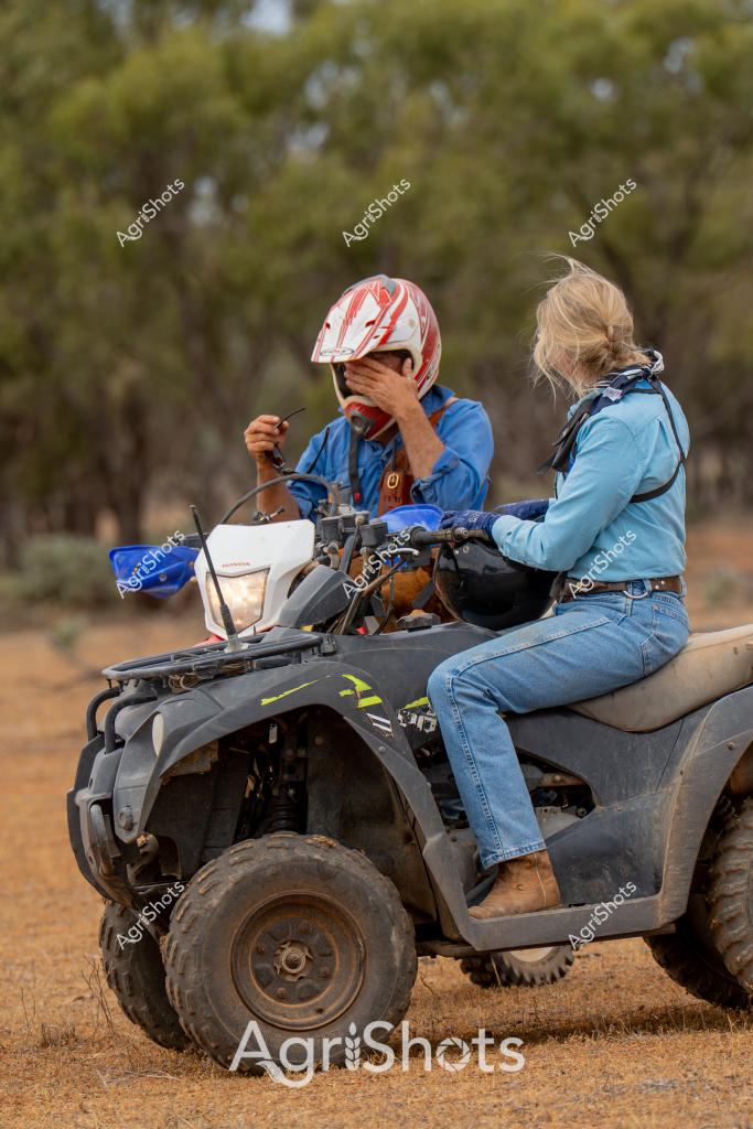 A woman and a young person sit on an all-terrain vehicle (ATV) in a dry, bushland setting, surrounded by sparse trees and dusty ground. The woman, wearing blue denim and a light blue jacket, is seated behind the rider, who is wearing a red and white helmet and a blue top. The grey ATV appears well-used, with robust off-road tires suitable for rugged rural terrain. The rider seems momentarily paused, with one hand covering their face, suggesting a brief moment of reflection or rest during what appears to be a farm or station work activity. The surrounding landscape looks typical of Australian rural areas - sparse eucalyptus trees, reddish-brown earth, and an open, expansive feel that characterizes many agricultural regions. The image captures an authentic slice of rural life, highlighting the practical use of all-terrain vehicles in managing large properties or conducting agricultural work.