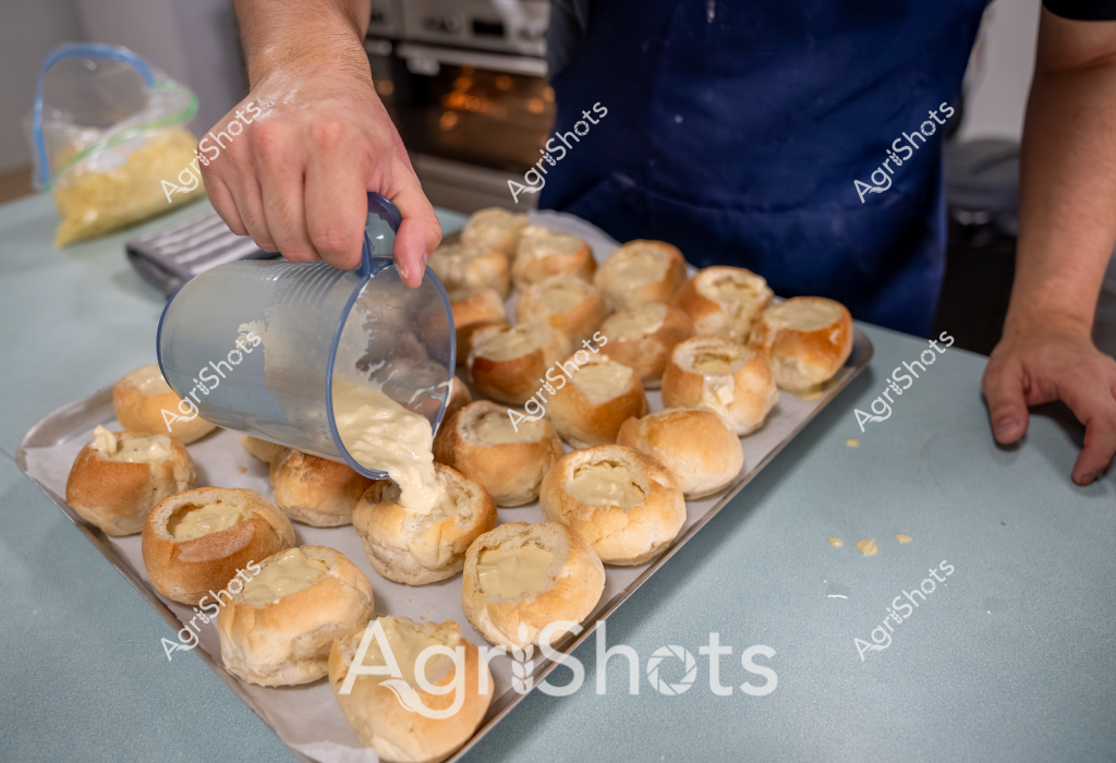 A person in a blue apron is preparing a batch of profiteroles or cream puffs in a kitchen setting. Using a clear measuring jug, they are carefully filling the golden-brown, hollowed pastry shells with a creamy filling. The delicate choux pastry buns are arranged neatly on a baking tray, showcasing a precise and methodical approach to pastry preparation. The soft, buttery color of the pastries contrasts with the blue of the baker's apron, creating a visually appealing scene of culinary craftsmanship. The image captures the delicate process of filling these classic French pastries, highlighting the skill and attention to detail required in baking.