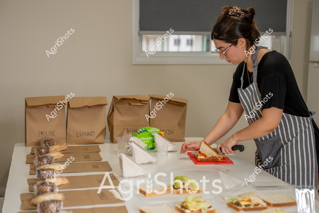 A woman wearing a striped apron and glasses prepares sandwiches in what appears to be a home kitchen or food preparation area. Multiple brown paper lunch bags are labeled with names like "Pauline", "Lennart", and another partially visible name, suggesting a catering or meal delivery service. She is working on a cutting board, carefully assembling sandwiches on fresh bread with various ingredients. The workspace is neat and organized, with packaged ingredients and pre-portioned items arranged systematically across the counter. The scene conveys a sense of methodical food preparation, with attention to detail in packaging and presentation. The soft, neutral background and window with a roller blind create a calm, professional atmosphere for what seems to be a small-scale food service operation.
