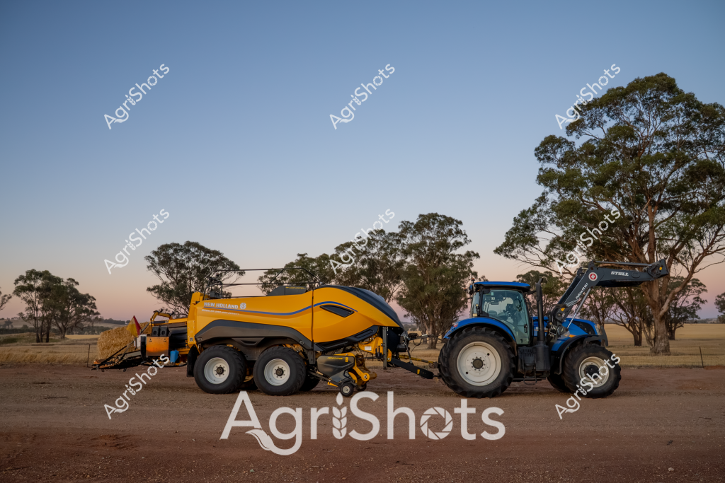 A New Holland tractor and large square baler stand ready in a dry, dusty rural paddock during the golden hour, capturing the essence of Australian agricultural harvesting. The bright blue tractor with a front-end loader is paired with a vibrant yellow and blue square baler, both positioned against a backdrop of mature eucalyptus trees and an expansive, arid landscape. The late afternoon light casts long shadows across the parched ground, highlighting the sophisticated farming equipment used in modern hay and crop production. This scene represents the precision and scale of contemporary agricultural machinery in Australian farming, where advanced equipment enables efficient harvesting even in challenging environmental conditions. The stark contrast between the modern, gleaming machinery and the rugged, sun-baked landscape epitomizes the resilience of Australian agricultural practices.