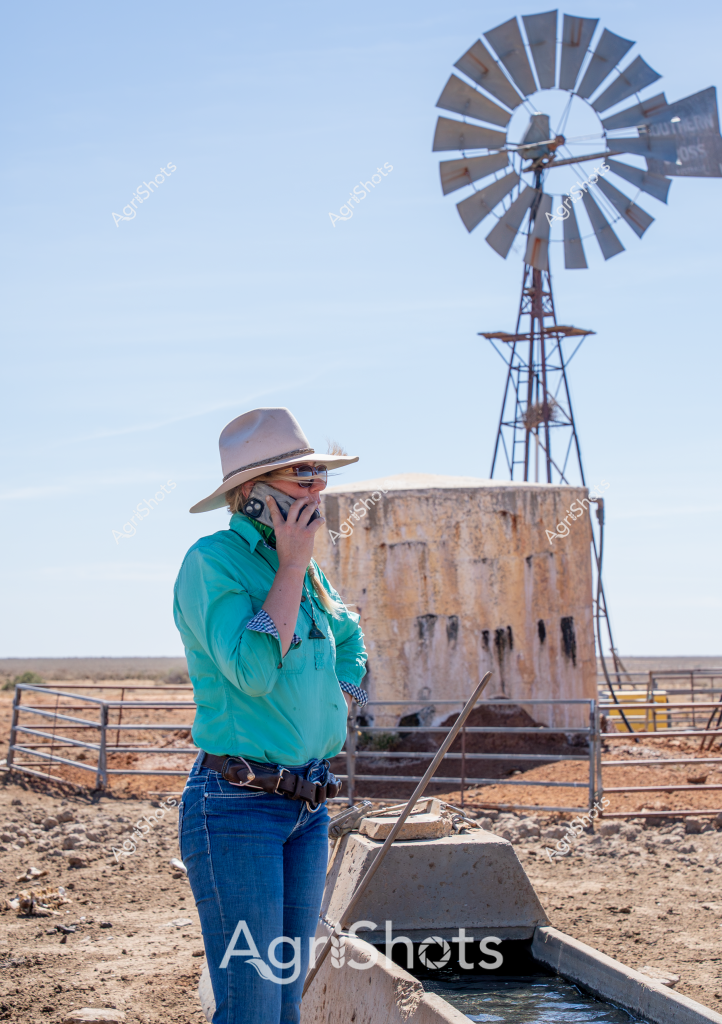 A woman farmer stands in a sun-bleached rural landscape, talking on a mobile phone beside a classic Australian windmill and water tank. Wearing a cream Akubra hat, turquoise long-sleeved shirt, and blue jeans, she appears to be managing a remote pastoral property. The metal windmill towers above a concrete water trough, with a rusted water storage tank visible in the background. The surrounding terrain is arid and rocky, typical of Australia's inland agricultural regions, with a clear blue sky suggesting a hot, dry day. Her stance and communication suggest she is coordinating farm operations or checking on livestock in this challenging outback environment.

The scene captures the essence of contemporary rural Australian agriculture - a blend of traditional infrastructure like the windmill and modern communication technology, with a resilient farmer adapting to the demands of managing agricultural land in a harsh landscape.