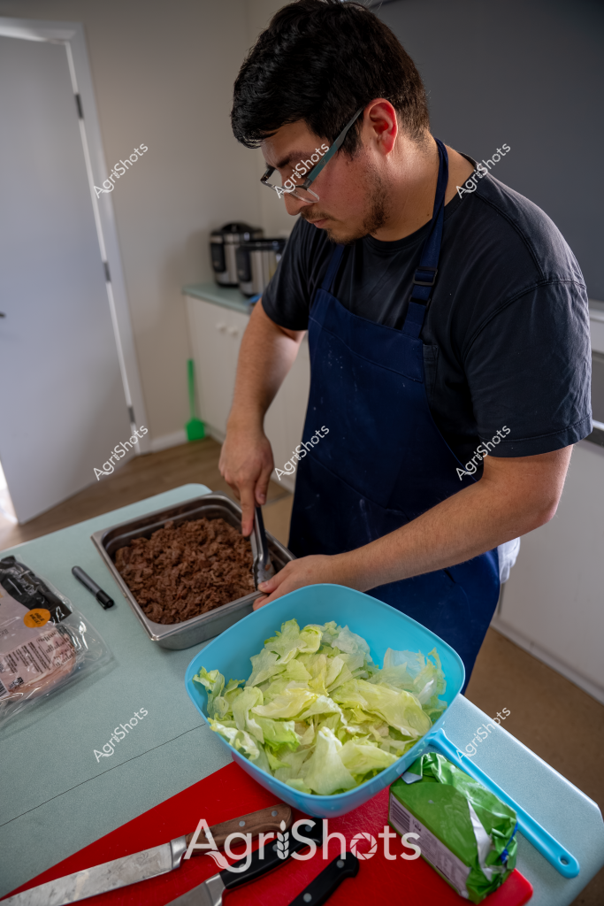 A man in a black t-shirt and blue apron prepares ingredients in a kitchen, focused on his culinary task. On the countertop, he has a blue bowl filled with chopped lettuce and a metal tray containing what appears to be ground meat. The kitchen setting is clean and bright, with a small appliance visible in the background and a cutting board with a knife in the foreground.

The man is wearing glasses and concentrating carefully as he works, suggesting he is methodically preparing a meal. The fresh ingredients and clean workspace indicate a home cooking scenario, with crisp lettuce and seasoned ground meat ready for preparation. His blue apron indicates a serious approach to cooking, whether he is preparing a home meal or practicing culinary skills.

The image captures a moment of food preparation, highlighting the careful attention and precision involved in cooking. The neutral kitchen environment and the man's focused demeanor create a sense of calm and intentionality in the kitchen workspace.