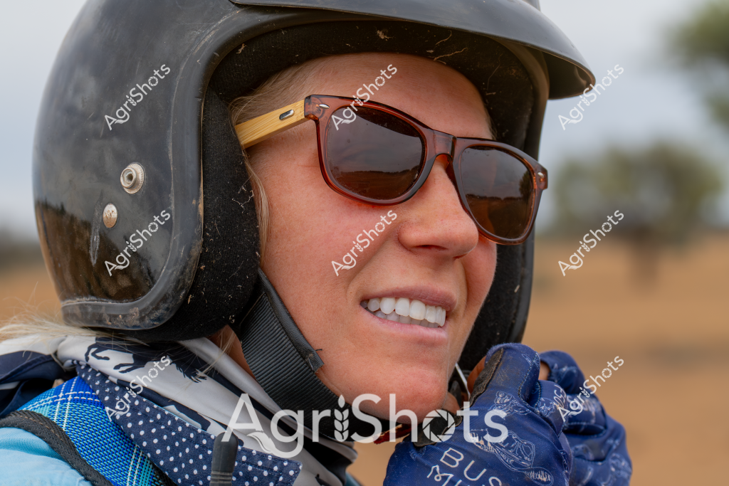 A woman wearing a black helmet and brown sunglasses smiles confidently while dressed in blue protective gear. Her warm expression and professional attire suggest she is engaged in an agricultural or outdoor working environment. The black helmet appears robust and well-fitted, indicating she may be involved in activities requiring safety equipment such as farm machinery operation, livestock management, or rural field work. Her blue gloves and patterned jacket further reinforce her preparedness for an active, hands-on role in agricultural settings. The background hints at an open, rural landscape with soft, muted tones typical of Australian farmland or station country.