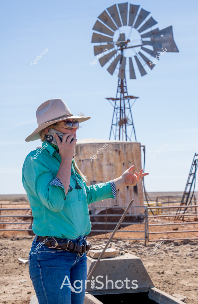 A woman wearing a broad-brimmed hat and teal work shirt stands in a dry, arid landscape beside a classic windmill and water tank. She is talking on a mobile phone while gesturing with her other hand, her stance conveying a sense of focused communication amid the stark rural setting. The windmill's metal blades stand silhouetted against a clear blue sky, while a concrete water tank and sparse fencing suggest this is a remote agricultural property. Her outfit - a crisp turquoise shirt, blue jeans, and a neutral-toned hat - reflects typical Australian rural workwear, highlighting the resilience and practical nature of farmers working in challenging outback conditions. The scene captures the essence of modern agricultural communication, where technology meets traditional farming landscapes.