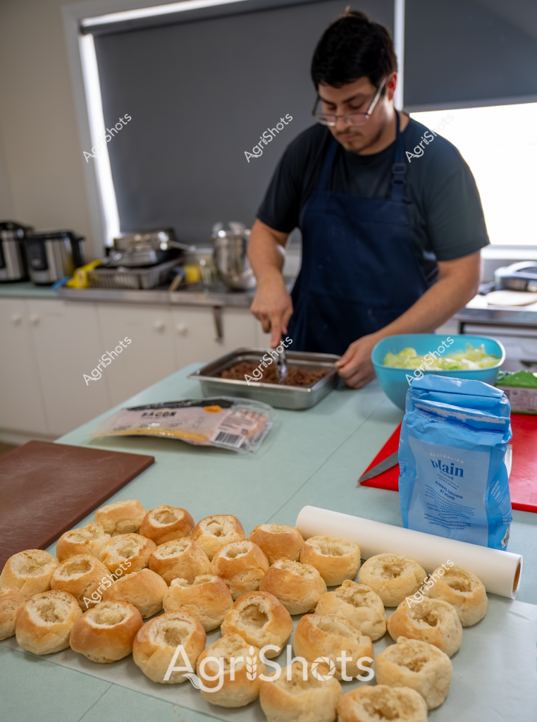 A professional kitchen scene captures a man in a navy apron preparing a batch of freshly baked bread rolls. Numerous golden-brown bread rolls are arranged neatly on the pale green work surface, their hollowed centers waiting to be filled. A blue bag of plain flour and various kitchen equipment are visible in the background, suggesting a professional food preparation environment. The man is focused on his task, carefully working with what appears to be a meat or filling tray, indicating he's in the process of creating filled bread rolls or a similar bakery product. The kitchen has a clean, organized appearance with stainless steel equipment and neutral-toned surfaces, typical of a commercial food preparation area.