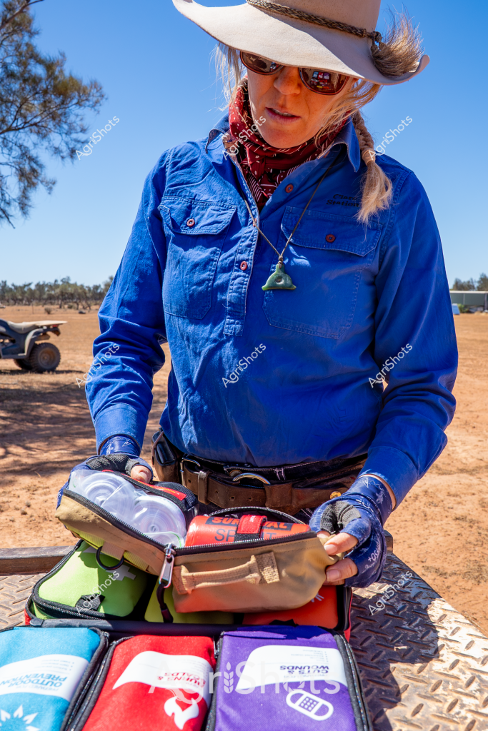 A woman agricultural worker stands in a dusty, arid rural setting, organizing medical or first aid kits on the tray of an all-terrain vehicle. Wearing a crisp blue long-sleeved work shirt, white wide-brimmed hat, and protective sunglasses, she carefully arranges multiple colored pouches and cases. A red bandana is tied around her neck, providing additional sun protection in the harsh outback environment. The metal vehicle tray beneath her has a textured surface, and an ATV is partially visible in the background, suggesting she is preparing equipment for remote work or emergency response in a challenging agricultural landscape. Her careful, methodical approach to organizing the kits indicates the importance of being prepared in isolated rural locations where quick medical access might be limited.